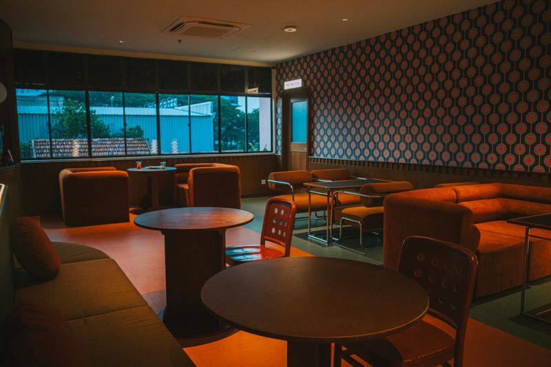 A retro-style lounge area with mid-century modern furniture, geometric wallpaper, and ambient lighting. The interior features a mix of cozy sofas, round tables, and soft textures, creating a relaxed environment ideal for socializing or unwinding in a stylish setting.