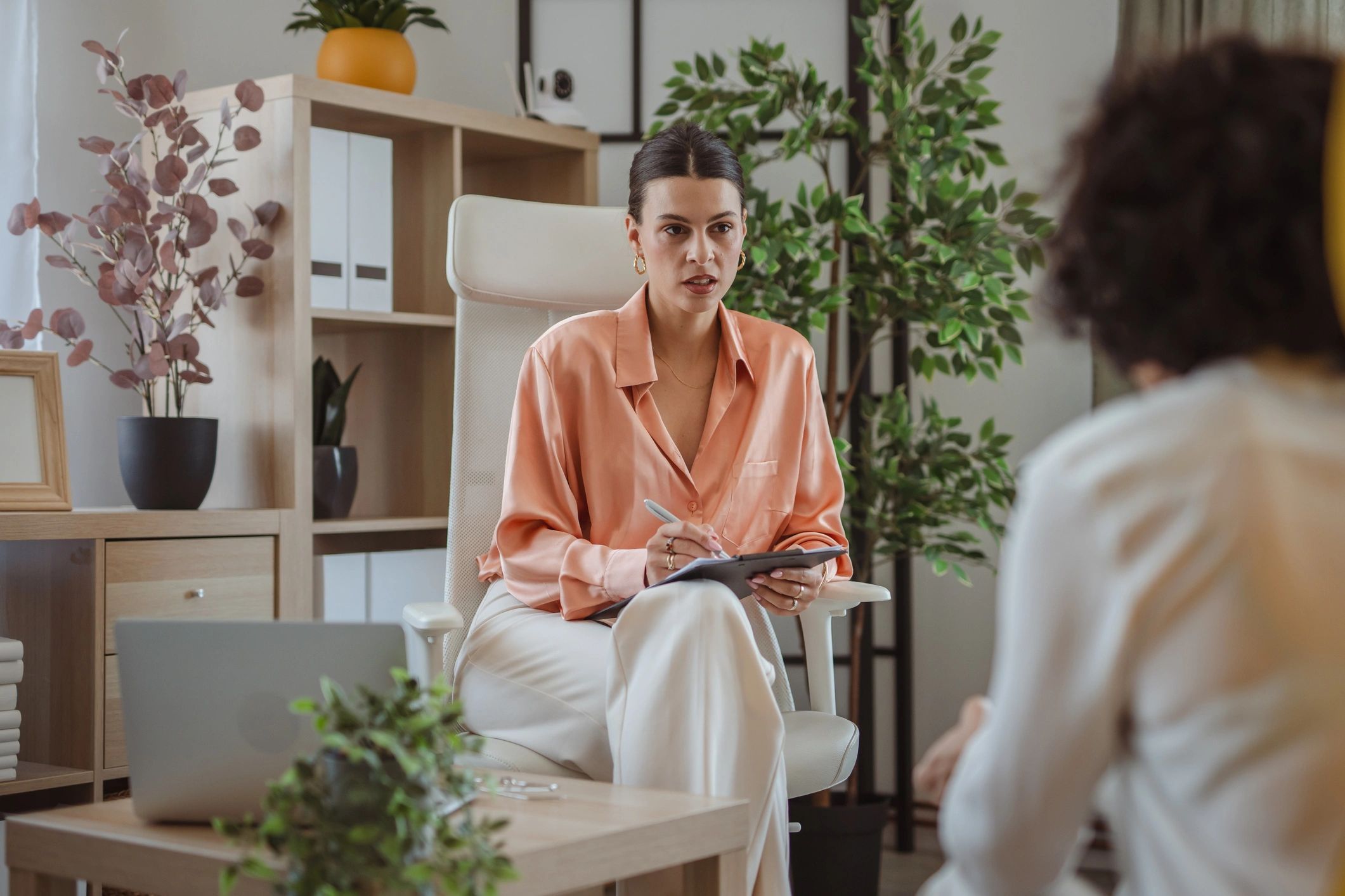 Therapist in a peach blouse seated in a white chair with a pen and tablet in a plant-filled office