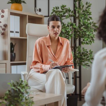 A woman in a peach blouse taking notes during a conversation.