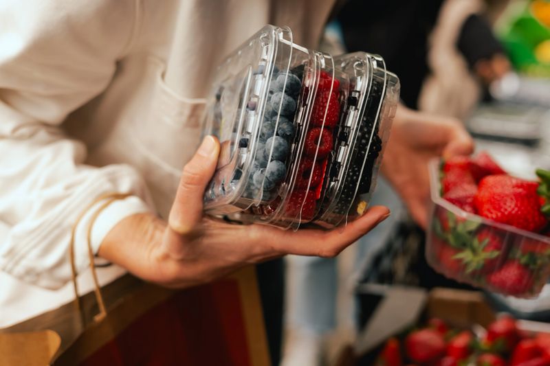 Close-up of hand with fresh berry in plastic packaging at local urban market. Organic produce on sale at outdoor farmer market. Selling fresh crops and veggies harvest. Part of the series