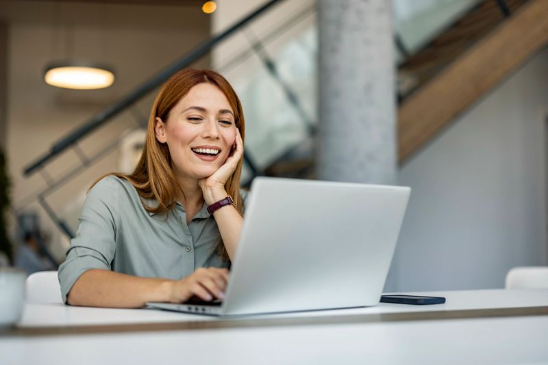 A smiling woman is working on her laptop while sitting at a desk in a modern office environment. The image conveys feelings of joy and productivity.