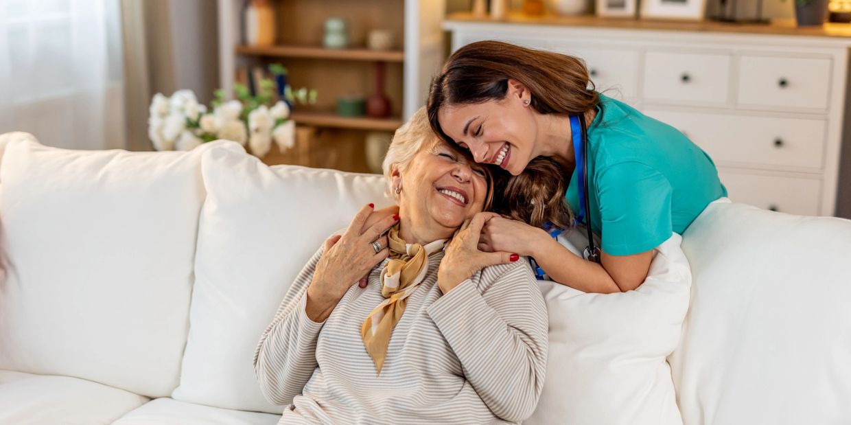 Caregiver and elderly woman sharing a joyful moment on a white sofa.