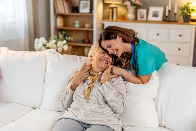 A compassionate nurse shares a warm moment with a smiling elderly woman, symbolizing care and companionship, in a cozy, sunlit living room, highlighting the importance of empathetic healthcare and well-being.