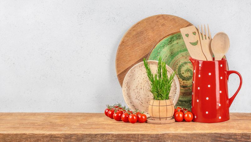 A cozy rustic kitchen setup with wooden utensils in a red polka dot jug, ceramic plates, cherry tomatoes, and fresh herbs arranged on a wooden counter against a light background.