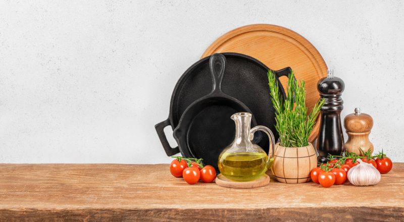 rustic kitchen counter scene with cast iron pans, fresh cherry tomatoes, olive oil, herbs, and pepper mills arranged neatly against a light wall backdrop.