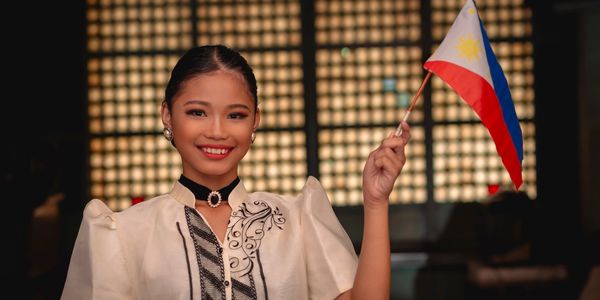 A smiling young woman in traditional Filipino attire holding a Philippine flag.