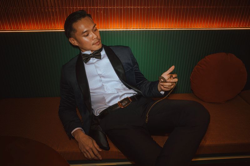 A fashionable young Asian man in a formal tuxedo and bow tie lounges on a modern sofa, gazing thoughtfully at a pocket watch. The warm ambient lighting and his composed demeanor evoke themes of elegance, timelessness, and refined masculinity. Perfect for concepts related to luxury lifestyle, time, fashion, and sophistication.