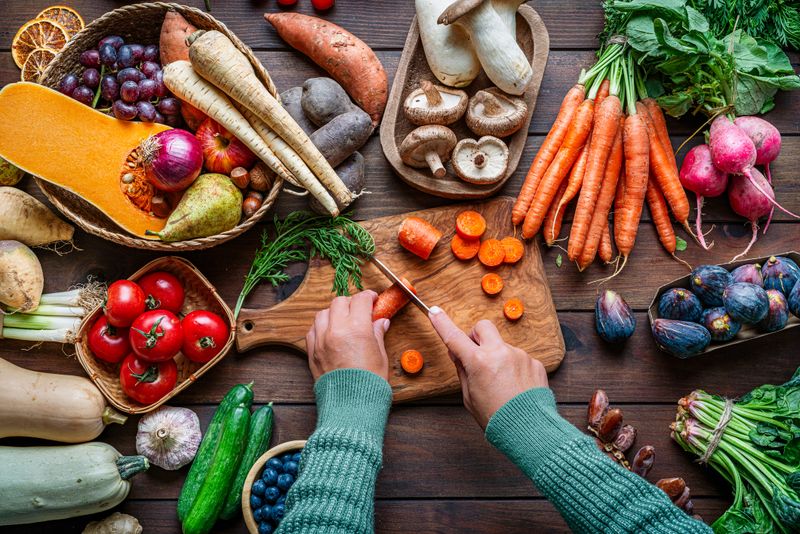 Woman chopping carrots on a table full of healthy Autumnal fruits and vegetables. Fresh organic seasonal produce background. Healthy vegetarian raw food. Food that boost the body inmune system. Nutrition and wellness, healthy diet and vegetable choice. Overhead view