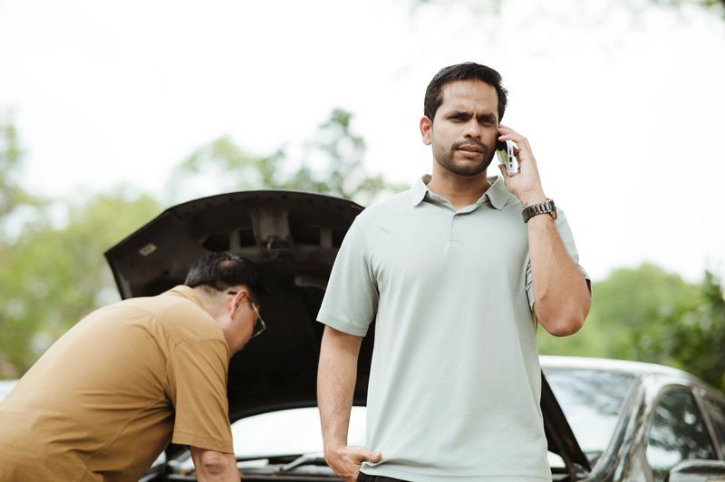 A mature man stands at the roadside, visibly stressed as he makes an urgent phone call for help following a sudden car breakdown. His friend, in the background, is bent over the open hood, trying to diagnose the issue. The scene captures a common travel emergency—unexpected mechanical failure, frustration, and the hope that help is on the way. The contrast between the man's anxious expression and his friend's hands-on troubleshooting reflects the shared tension of the moment.