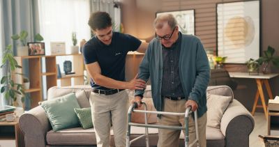 Young man helping elderly man with a walker inside a cozy living room.