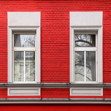Two white-framed windows on a vibrant red brick wall with tree reflections.