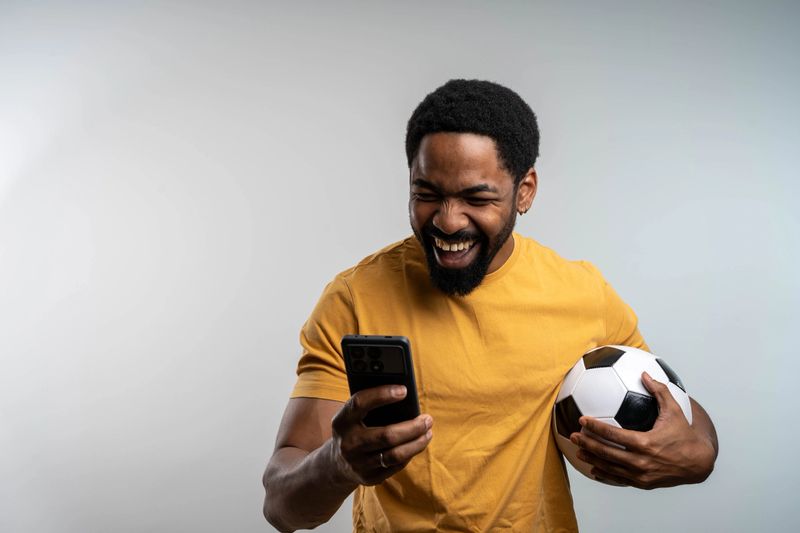 Excited African man celebrating win, holding smartphone in hand, happy euphoria mood, isolated studio background