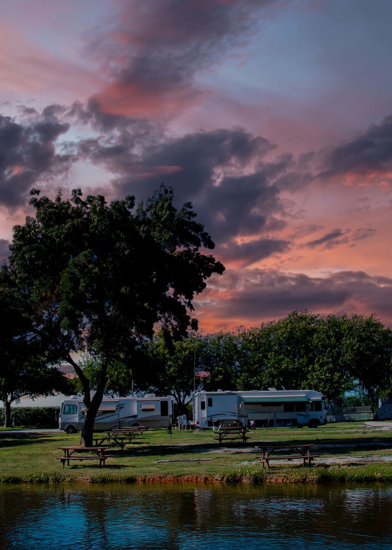 Recreational vehicles are parked near a tranquil pond, with picnic tables and trees dotting the grassy area. The sky is filled with dramatic clouds, painted in hues of pink and purple as the sun sets.