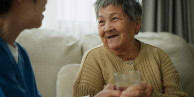 Oudere vrouw praat met verzorger en houdt een glas vast.