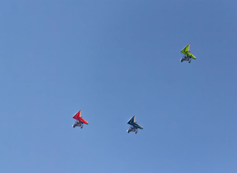 Three colorful ultralight aircraft, one red, one blue/green, and one yellow/green, soar through a vast, clear blue sky.