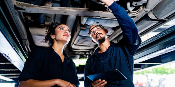 Two mechanics inspecting the underside of a vehicle in a garage.