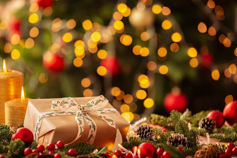 Christmas present wrapped with brown craft paper and a white ribbon on a cozy table with Christmas decore candle and illuminated tree at background. Gift or surprise, New Year's Eve. Copy space