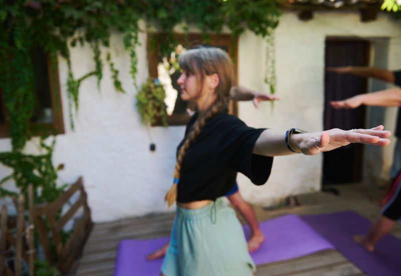 Women engaging in yoga poses on a wooden deck amidst greenery, emphasizing harmony and physical wellbeing during a serene outdoor yoga session in a tranquil garden setting.
