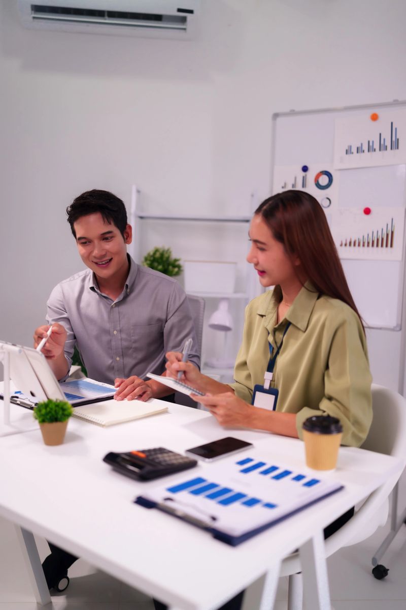 Two Asian adults discuss financial reports and charts in a bright office focusing on achieving company objectives and market expansion