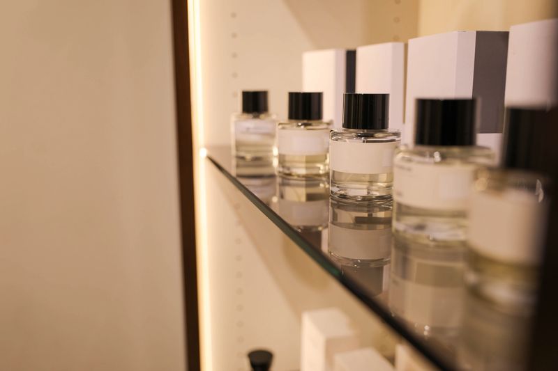 Close-Up Of Transparent Perfume Bottles With Black Caps Arranged Neatly On A Glass Shelf In A Well-Lit Retail Store. Minimalist Packaging And Soft Lighting Emphasize Luxury And Sophistication In Fragrance Shopping.