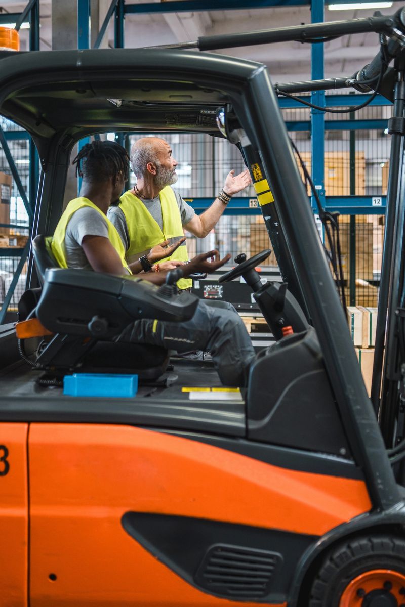 A young Black man and a mature Caucasian man, wearing yellow safety vests, work together operating a forklift in a brightly lit airport cargo warehouse with shelves and boxes in the background.