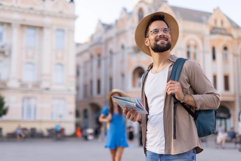 A bearded man with glasses and a stylish hat stands outdoors holding a map, with a European-style historic building as the background, suggesting exploration and adventure.