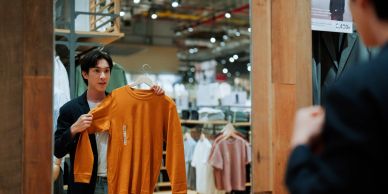 Man holding an orange sweater looking at himself in a store mirror.