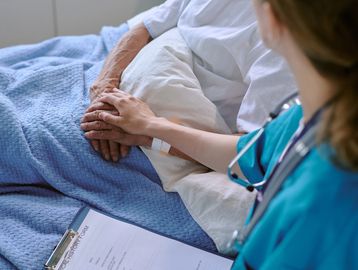 A nurse comforting an elderly patient by holding their hand in a hospital bed.