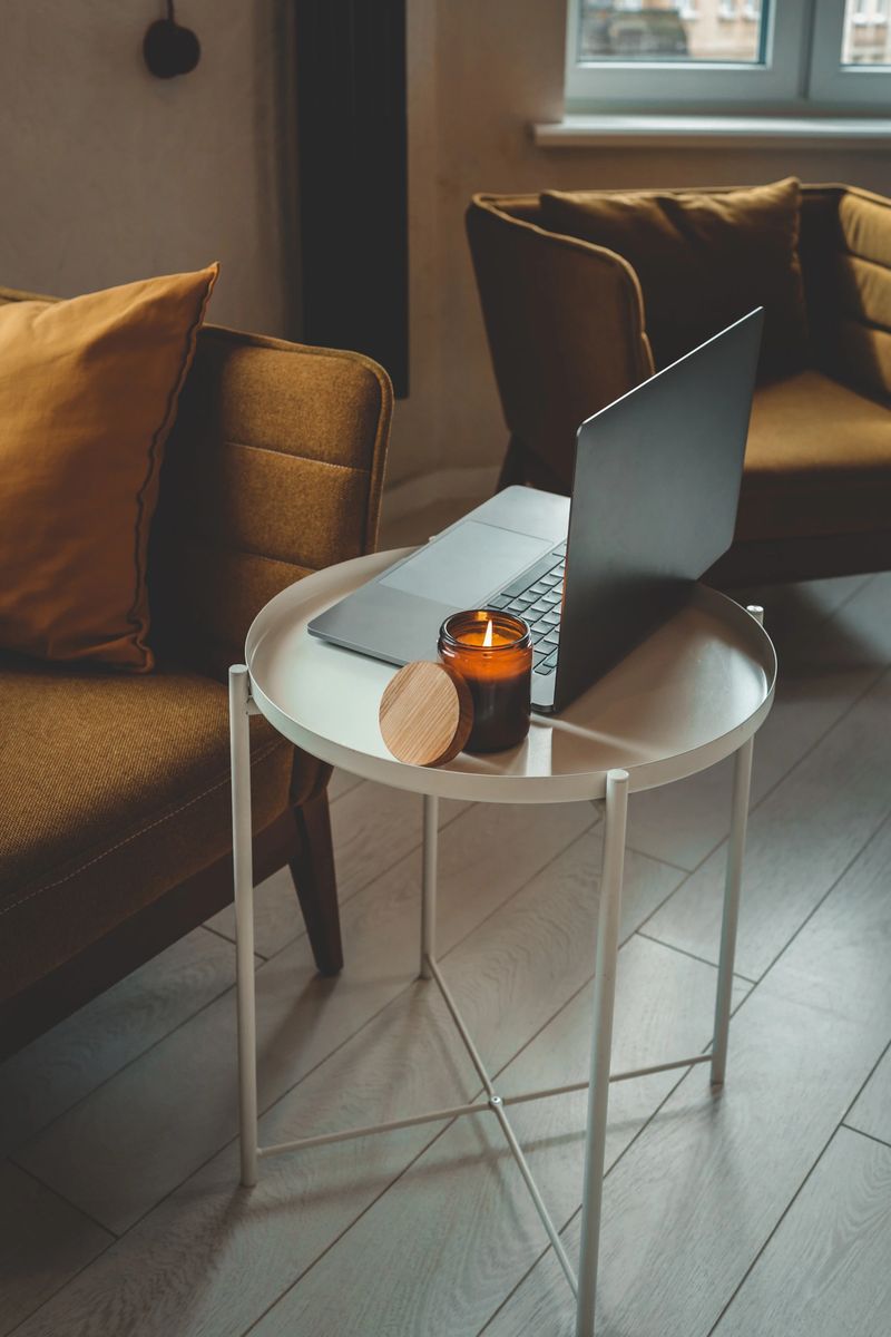 Cozy home interior with mustard armchair, burning candle in glass jar, and laptop on a small white table. Warm natural light and peaceful atmosphere.