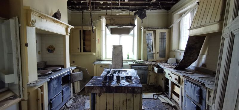 Eerie fire damaged home with sunlight through windows, island counter, aga oven cooker, burnt drawers and surfaces. Interior kitchen of derelict country mansion with signs of vandalism and arson. Destroyed room of domesitic residential building vandalised and burned remains from disaster. Haunted feel of natural light and shadow across white kitchen worktops contrasting blackened charred wood.