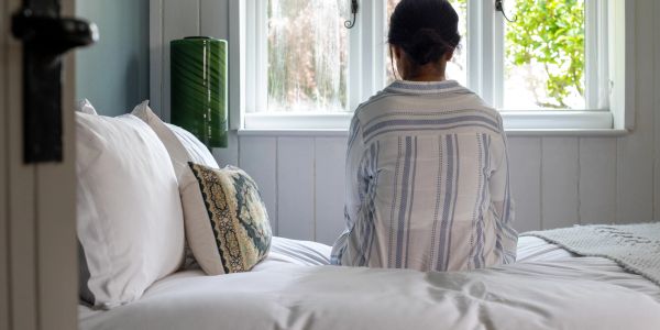 Person sitting on a bed facing a window in a cozy room.
