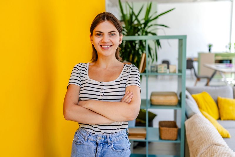 A young woman stands with arms crossed in a bright indoor setting featuring a yellow wall and stylish furniture. She exudes confidence in her casual attire while being surrounded by greenery.