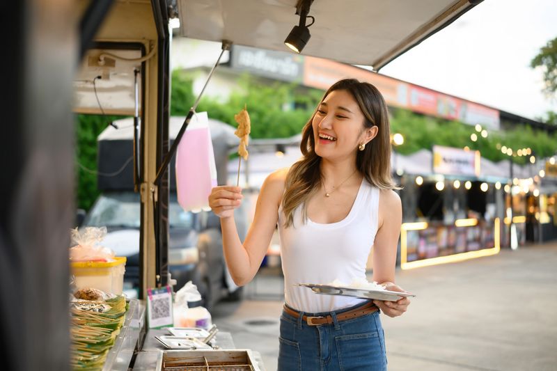 Young woman holding a plate and MALA BBQ skewer at a street food truck, enjoying authentic local flavors in an open-air market filled with lights and vibrant energy.