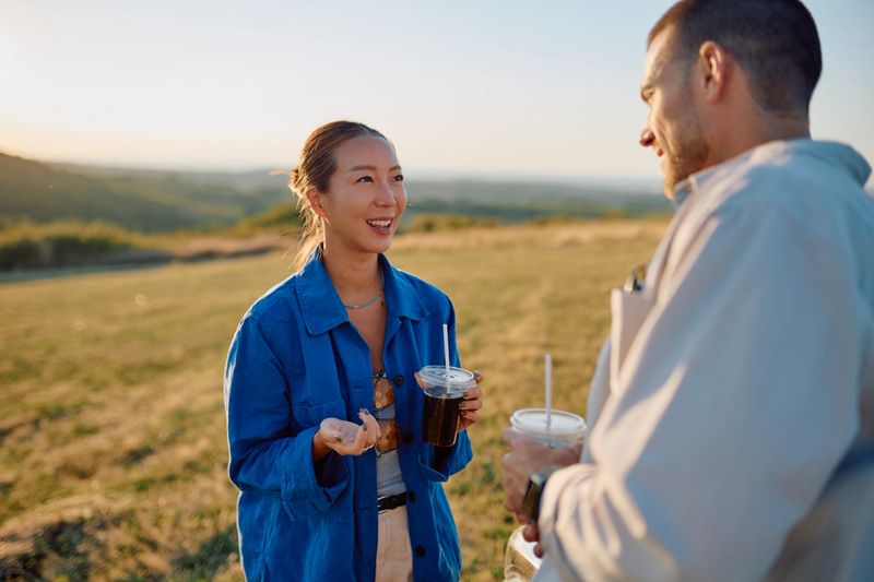 Young multi-ethnic couple enjoying coffee and chatting in a scenic field at sunset, soaking in the beautiful view and warm atmosphere