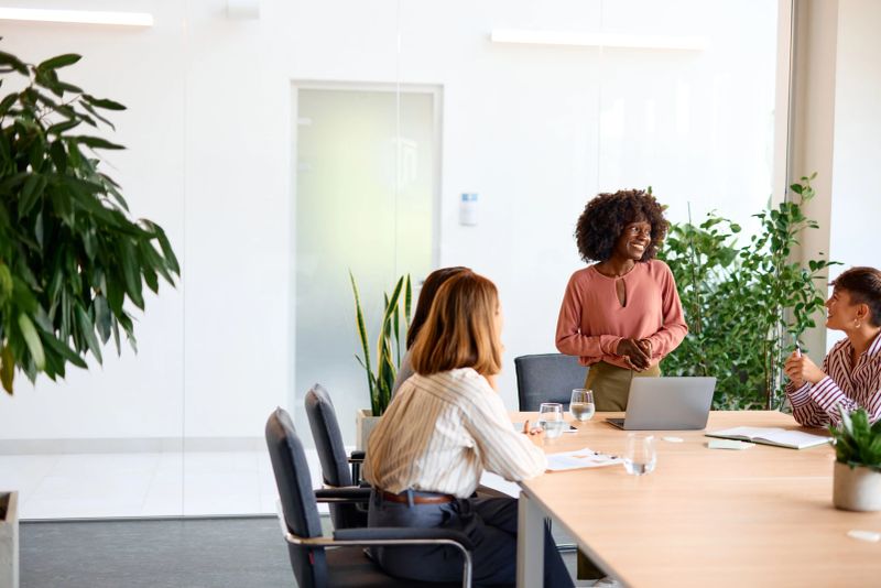 Four businesswomen are discussing a project in a modern office, sitting around a table with a laptop and documents, with plants adding a touch of green to the workspace