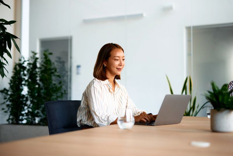 Young Asian businesswoman sitting at a modern office desk, working intently on her laptop. Focused expression reflects her dedication to achieving professional goals in a bright, clean workspace