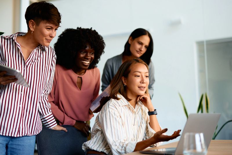 Four diverse businesswomen are gathered around a laptop, collaborating on a project in a modern office environment, with one woman presenting information on the screen