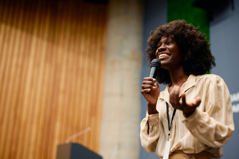 Young woman delivering an engaging speech while holding a microphone, smiling and gesturing confidently in a spacious auditorium filled with an attentive audience