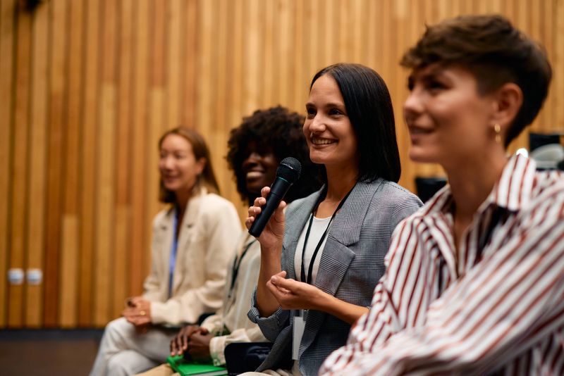 Group of businesswomen engaging at a conference, one speaking into a microphone while others are smiling and attentively listening, fostering an atmosphere of collaboration and inspiration