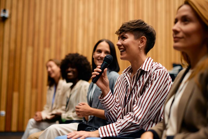 Businesswoman holding a microphone and speaking at a conference, engaging with a group of fellow businesswomen in a modern conference room, fostering discussion and collaboration