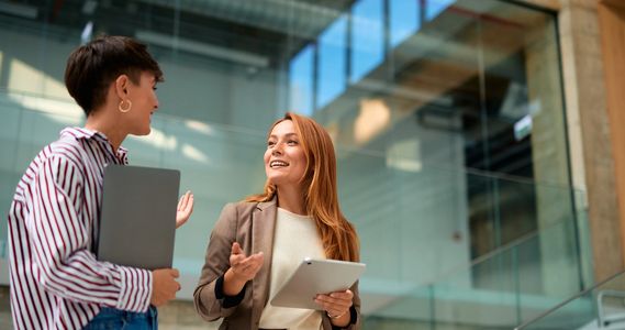 Two women in a modern office chatting with tablets in hand.