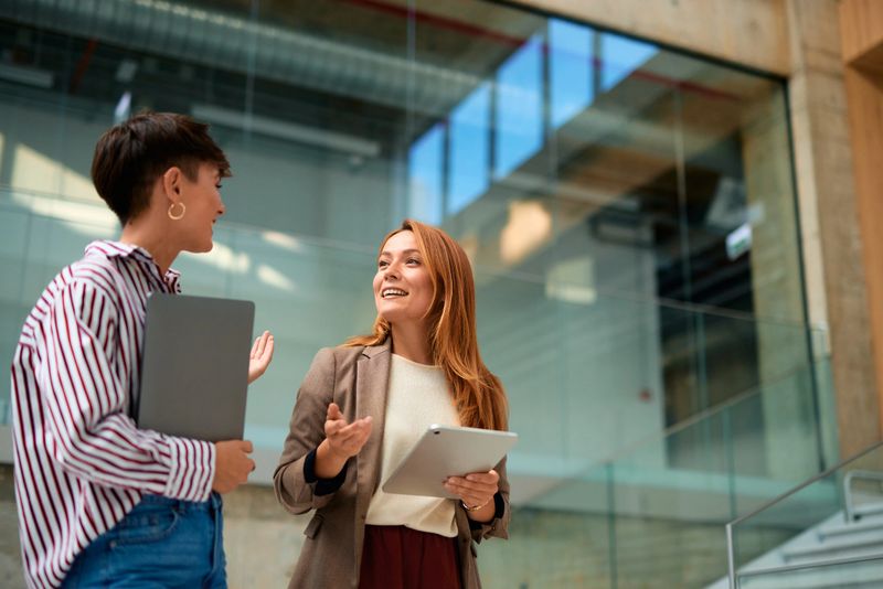 Two young businesswomen engaging in a project discussion while holding a laptop and a tablet, situated in a modern office building featuring large glass windows and elegant stairs