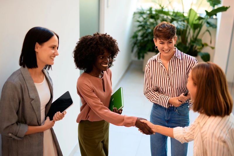 Four businesswomen shaking hands and smiling in an office hallway, exchanging greetings while embodying teamwork and collaboration in a professional environment
