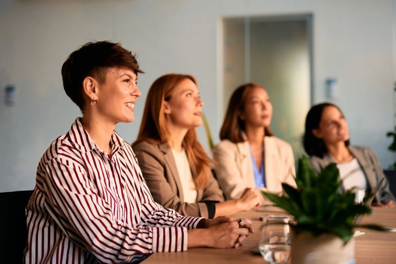 Four businesswomen sitting at a conference table, listening attentively to a speaker during a business meeting in a modern office, embodying teamwork and collaboration in a professional setting