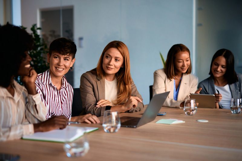 Five businesswomen sitting at a modern office table, engaging with a presentation and sharing smiles, exemplifying teamwork and collaboration during a productive workday