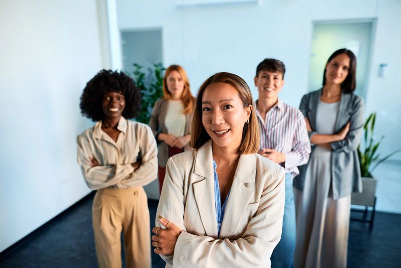 Five multi-ethnic businesswomen smiling with arms crossed, exuding confidence and showcasing teamwork in a modern office setting, embodying strength and empowerment in their professional roles