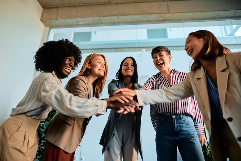 Five cheerful businesswomen joining hands in the office, celebrating a successful project together, embodying strong teamwork and unity while radiating joy and motivation