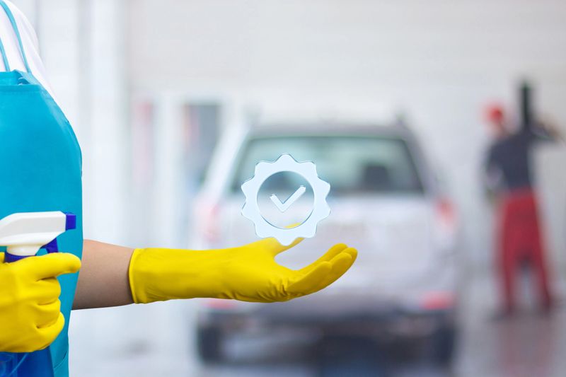 A worker in yellow gloves holds a spray bottle, ready to clean, while another employee details a vehicle in the background of a car wash. Bright, organized environment enhances efficiency.