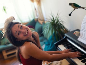 A woman joyfully plays piano with birds perched around her.