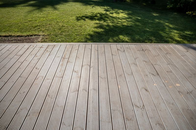 A wooden deck with visible grooves in the foreground, leading to a grass lawn. Tree shadows create contrast and texture on the well maintained grass.